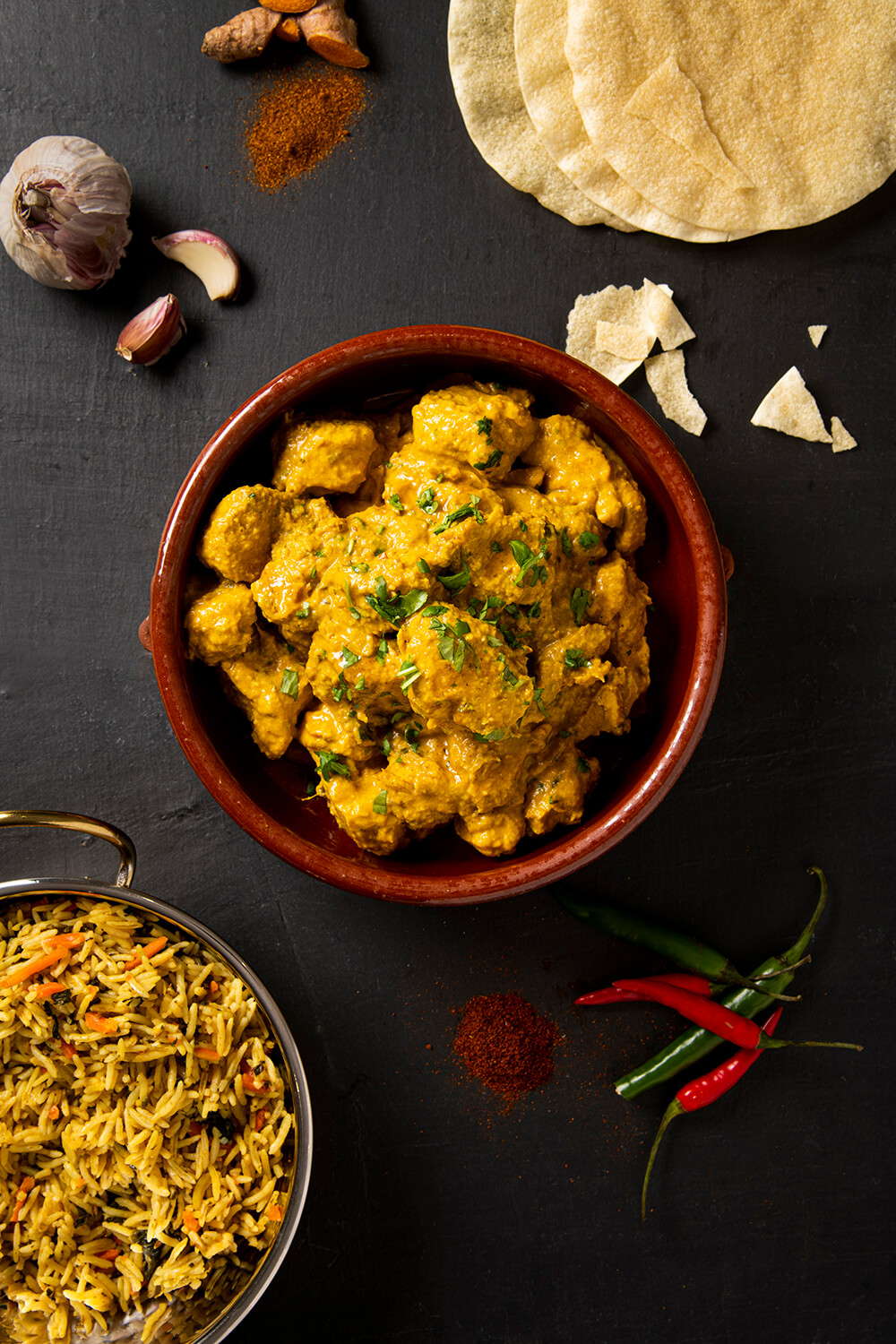 Food photography of chicken Korma taken from above. The shot has been styled to include props such as a bowl and ingredients such as chillies and popadoms and rice. They are sitting on a background of black slate. By Glasgow food photographer Richard Campbell
