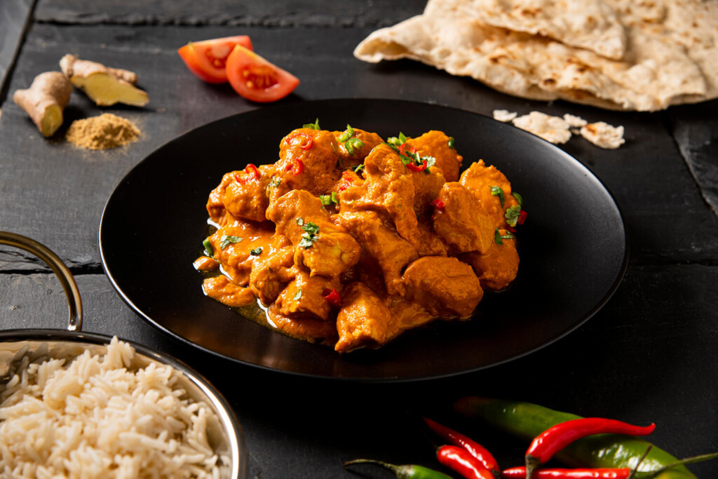 Food photography of chicken tikka taken from the side. The tikka is orange and is in a black bowl. You ca see other things nearby such as ginger, chillies, nan bread and rice. They are all on a background of black slate. By Glasgow food photographer Richard Campbell