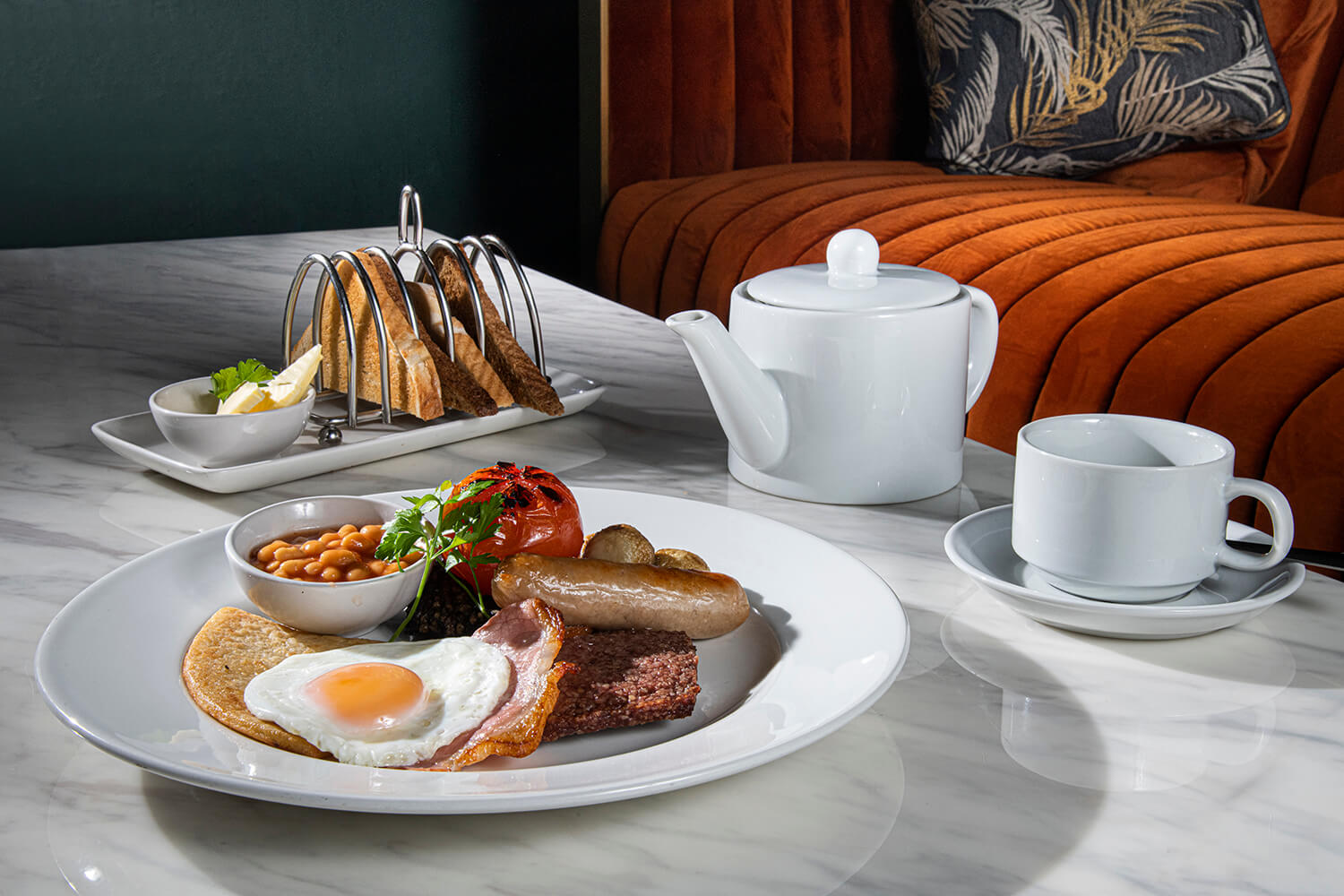 Food photography of cooked breakfast. There is a plate with bacon, egg, sausages and tomatoes in the foreground with a rack of toast, white teapot and white cup in the background. By Glasgow food and hospitality photographers Blink Imaging