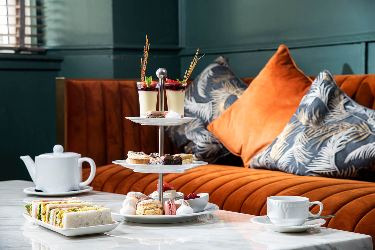 Food photography of an afternoon Tea. There is a cake stand sitting on a low marble table. Next to this is a white teacup and teapot with a plate of sandwiches. You can see an orange sofa and cushions in the background. By Glasgow hospitality and food photographers Blink Imaging