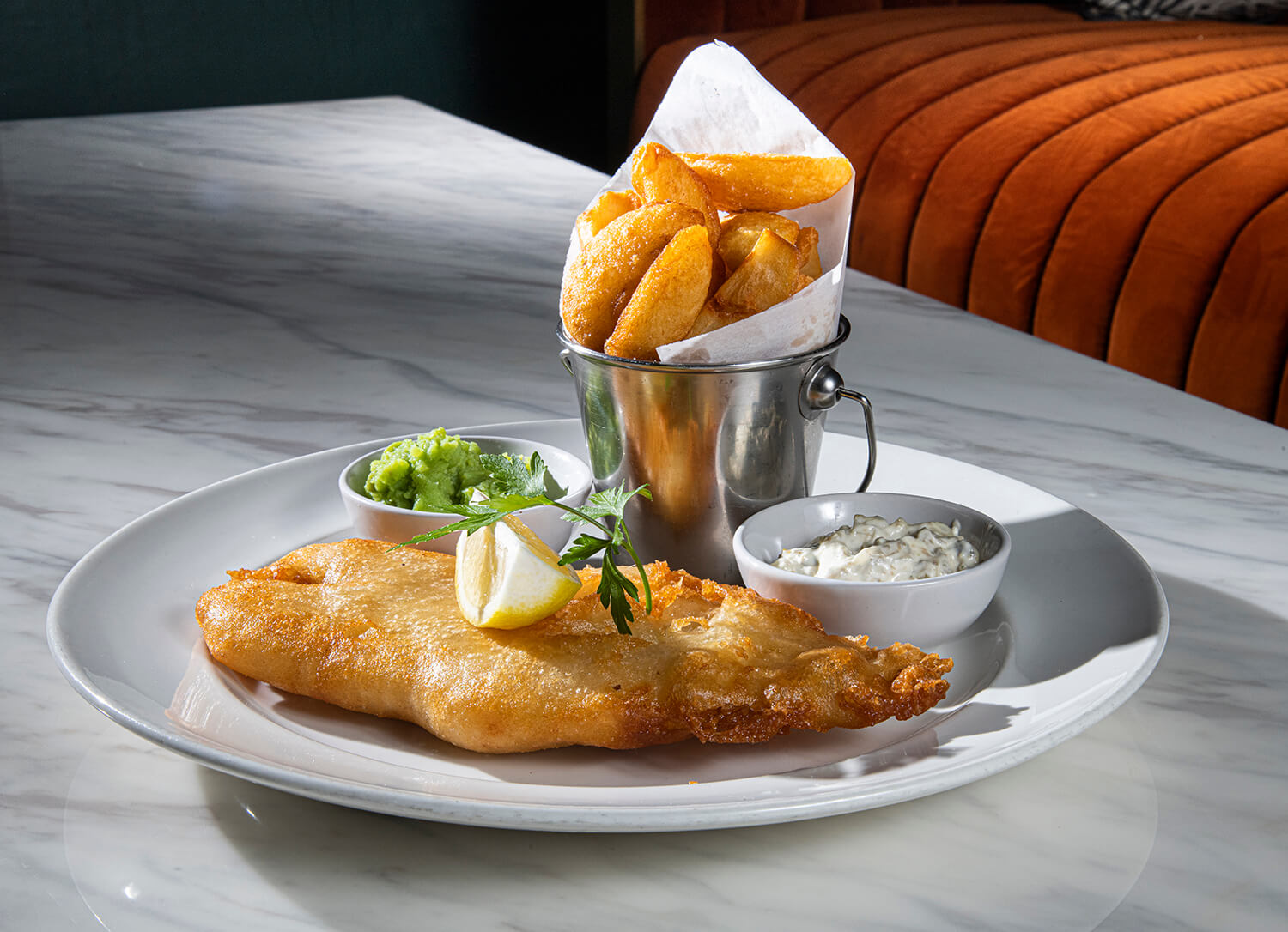 Food photography of fish and chips. They are on a white plate which is sitting on a marble table. The chips are sitting up in a metal container. There are also mushy peas on the plate. In the background you can see an orange sofa. By Glasgow hospitality and food photographers Blink Imaging