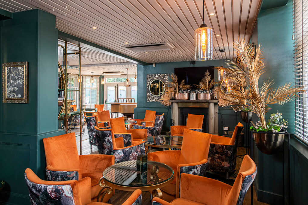 Interior of new hotel lounge at the Kirkhouse Inn. You can see a conservatory in the background with light streaming through. In the foreground you can see orange chairs. The walls are turquoise. There are old fashioned lights on the ceiling and artificial gold ferns in plant stands. By hospitality and leisure photographers Blink Imaging