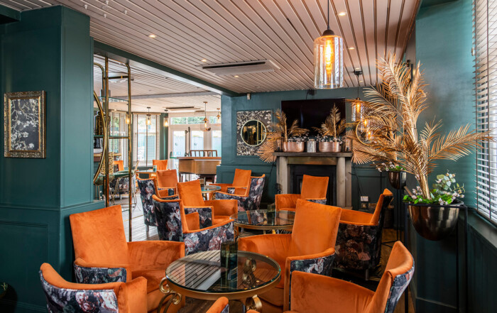 Interior of new hotel lounge at the Kirkhouse Inn. You can see a conservatory in the background with light streaming through. In the foreground you can see orange chairs. The walls are turquoise. There are old fashioned lights on the ceiling and artificial gold ferns in plant stands. By hospitality and leisure photographers Blink Imaging