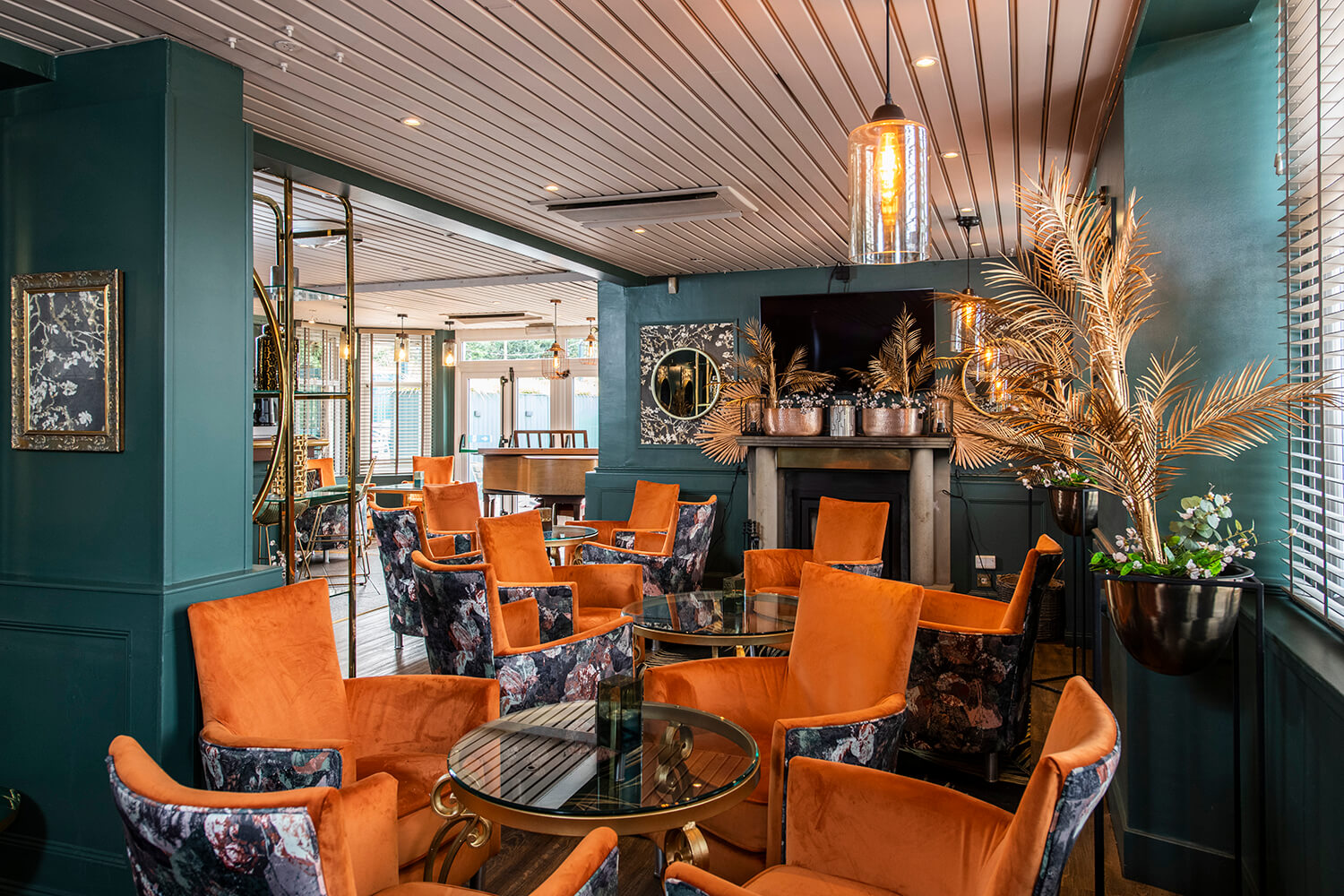 Interior of new hotel lounge at the Kirkhouse Inn. You can see a conservatory in the background with light streaming through. In the foreground you can see orange chairs. The walls are turquoise. There are old fashioned lights on the ceiling and artificial gold ferns in plant stands. By hospitality and leisure photographers Blink Imaging