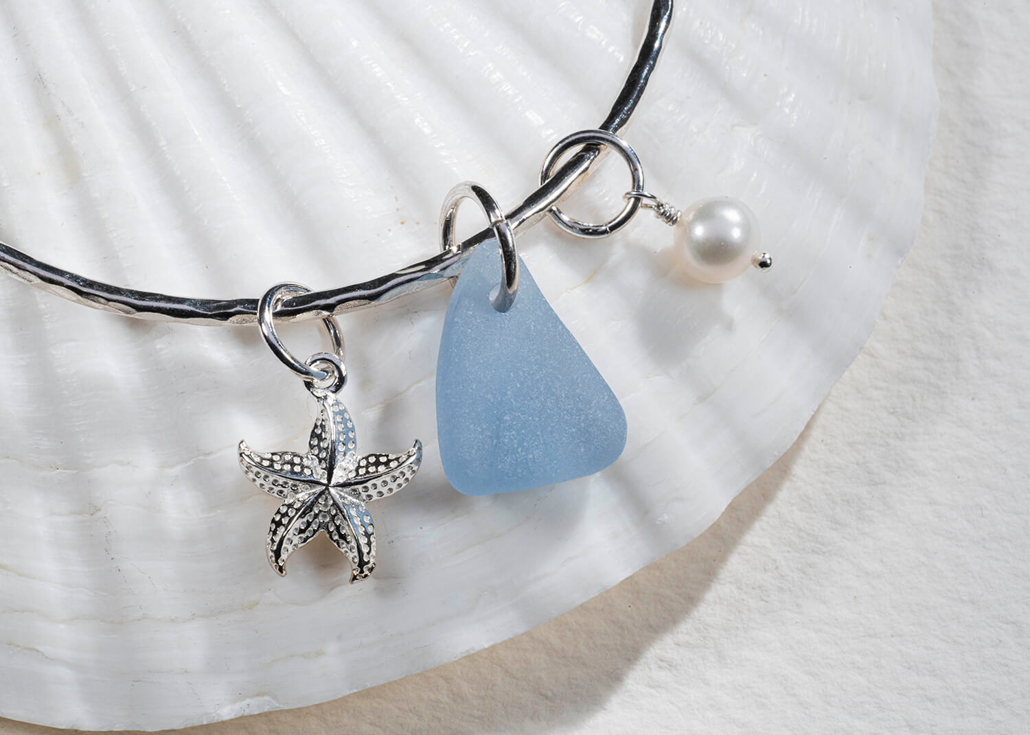Advertising photograph of sea glass jewellery sitting on sea shell. The sea glass is blue and is attached to a silver bracelet with some silver charms attached. By Glasgow jewellery photographers Blink Imaging