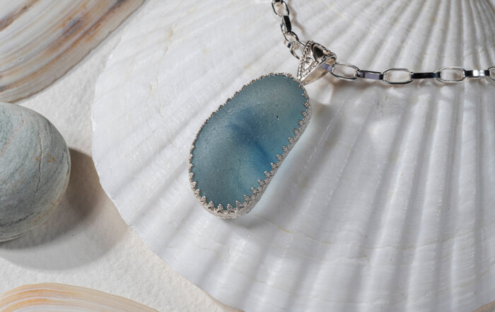 Product photo of seal glass jewellery. The blue sea glass is attached to a silver chain and is sitting on top of a sea shell. The light is shining through the glass and you can see other pebbles in the image by Glasgow jewellery photographers Blink Imaging