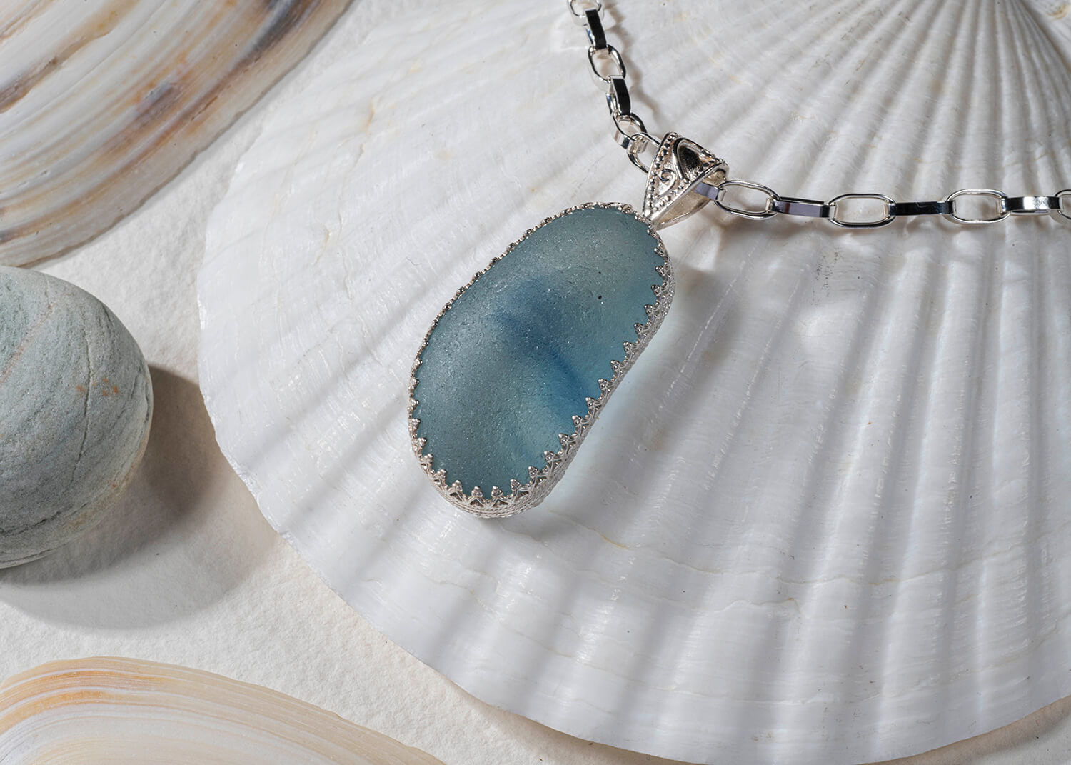 Product photo of seal glass jewellery. The blue sea glass is attached to a silver chain and is sitting on top of a sea shell. The light is shining through the glass and you can see other pebbles in the image by Glasgow jewellery photographers Blink Imaging