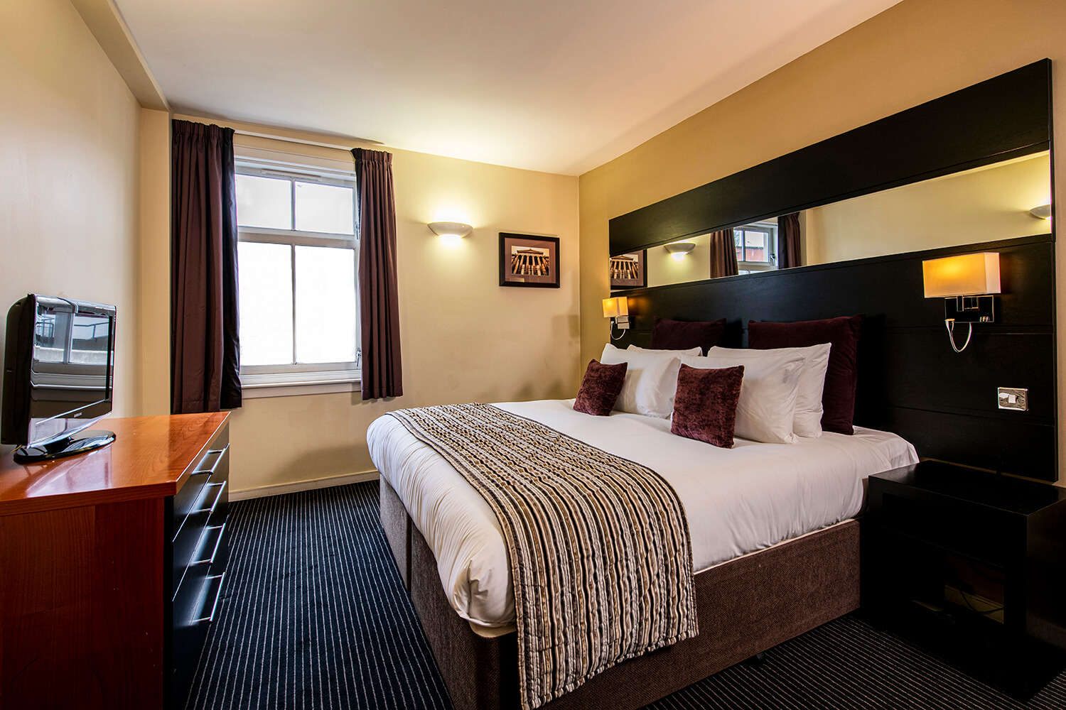 Interior photograph of bedroom. There is a double bed in the middle of the room with cushions on it. Above this is a large dark wooden headboard with lights and a long mirror. Opposite this is a set of drawers with a TV. There is light coming in the single window. By Glasgow interior photographers Blink Imaging