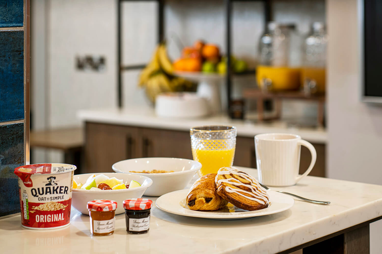 Close up of continental breakfast on breakfast bar. You can see pastries on a plate in the foreground. Next to this are bolts of fruit and cereal. There are also pots of jam, a pit of Quaker Oats, a glass of orange juice and a mug of tea. In the background you can see a self serve area with fruit and fruit juice dispensers.