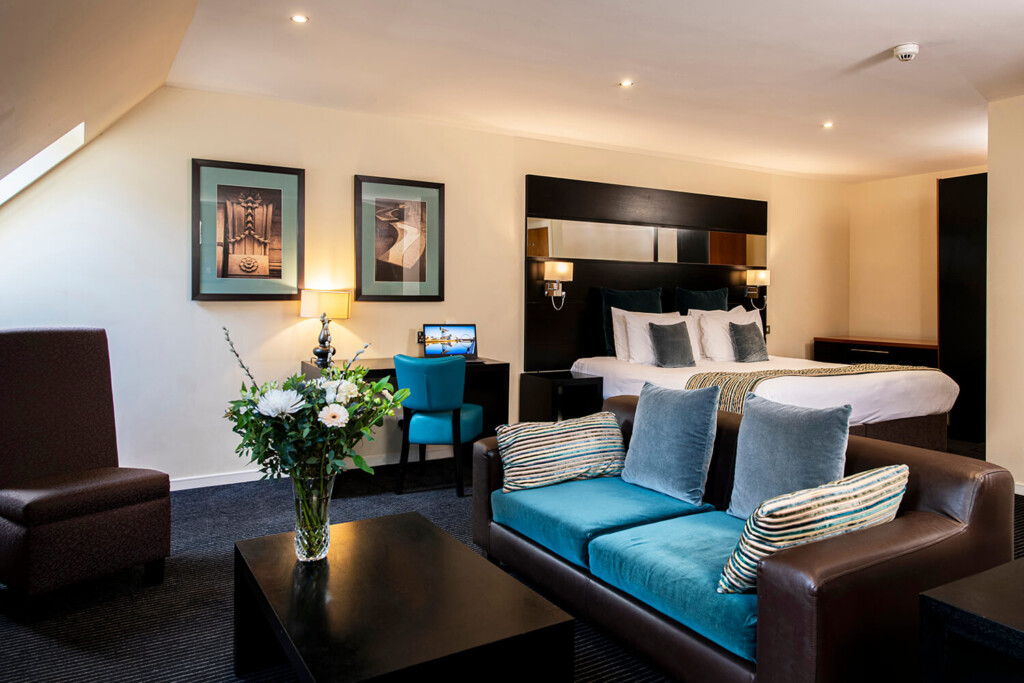 A view of the lounge and bedroom area of a serviced apartment. There is a brown leather sofa with blue cushions in the foreground. Behind this you can see a double bed and a desk and chair with a laptop. There are flowers on a table in front of the sofa. By Glasgow commercial interior photographers Blink Imaging