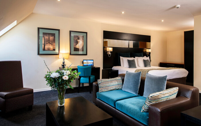 A view of the lounge and bedroom area of a serviced apartment. There is a brown leather sofa with blue cushions in the foreground. Behind this you can see a double bed and a desk and chair with a laptop. There are flowers on a table in front of the sofa. By Glasgow commercial interior photographers Blink Imaging