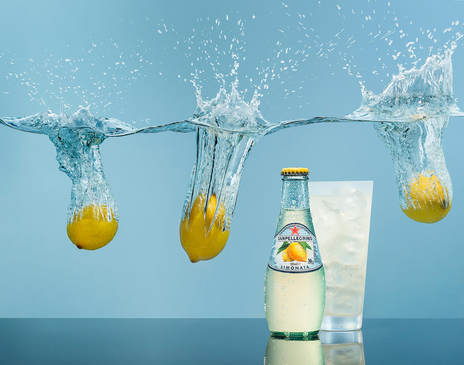 Branding photography for lemonade. There is a bottle of lemonade on the right next to a frosted glass with ice. Lemins can be seen splashing through the surface of water above them. By Glasgow photographers Blink Imaging